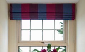 a Geogian style sash window with colourful blinds overhead and complementary red flowers in a modern chrome vase below, next to two brown candles. In the background is a bright view to a garden with lots of trees.